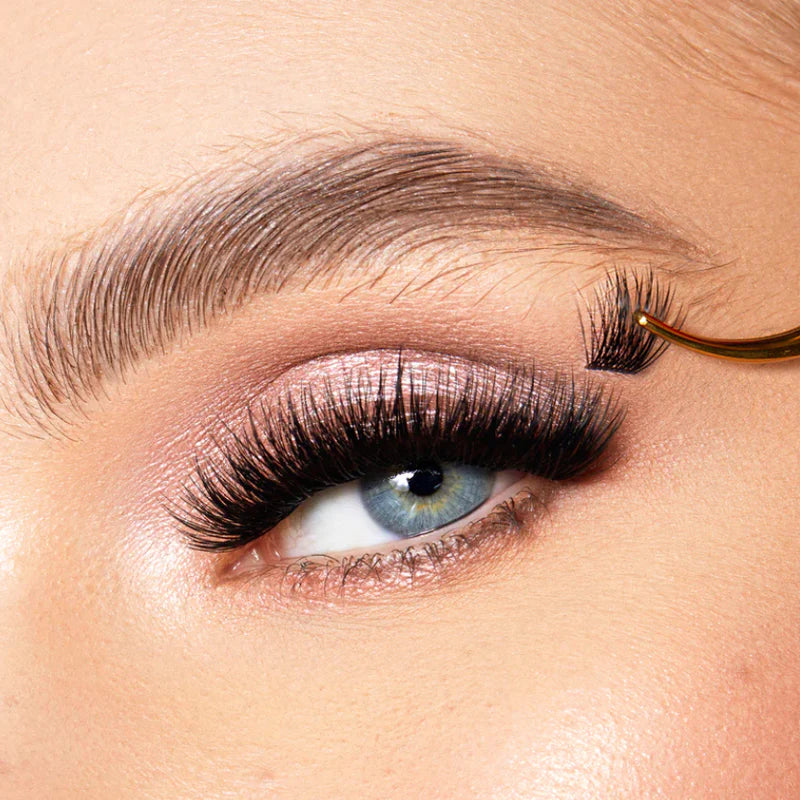 A close-up image of a person's eye with false lashes applied. The lashes are longer and thicker than natural lashes, and they curve upwards. The lashes are attached to the person's natural lashes using adhesive